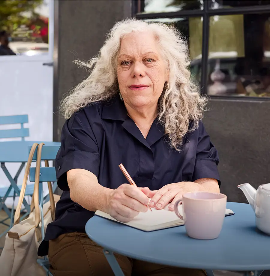 An older woman sitting at an outdoor cafe, writing in a journal.
